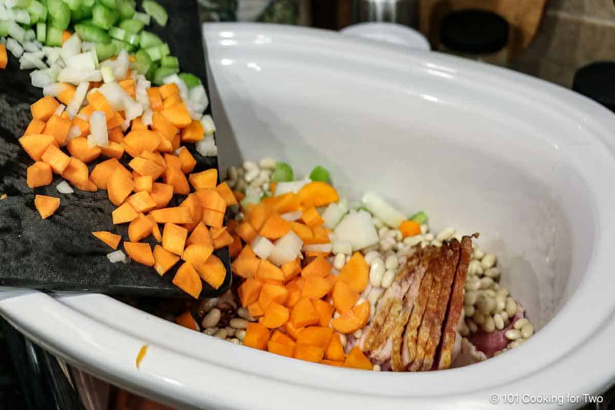 Pouring chopped veggies into crock pot with a ham bone.