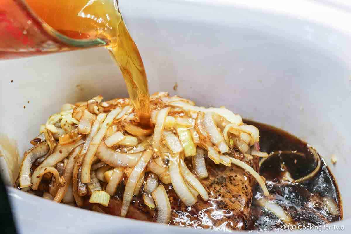 Pouring broth into the crock pot with beef and onions.